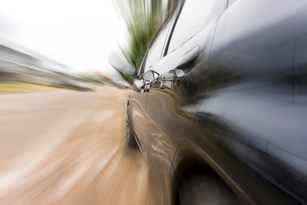 A dark car speeds down a road with a blurred motion effect showing high velocity and surroundings in a streaked background.