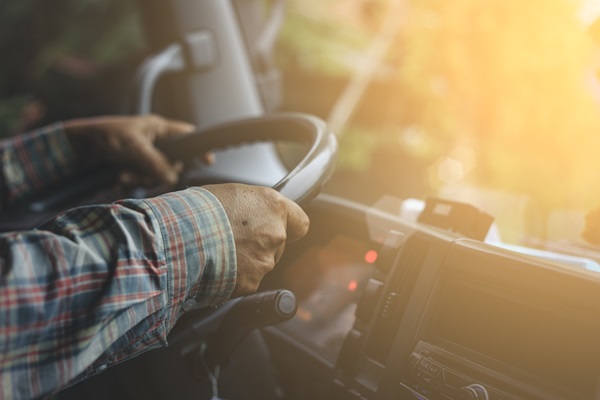 The hands of a male truck driver on the steering wheel of a semi-truck with sunlight streaming through the windshield.