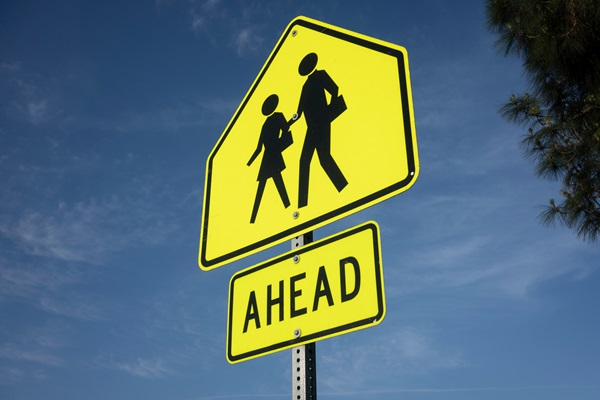 Bright yellow pedestrian school zone sign with the word “Ahead” against a blue sky.