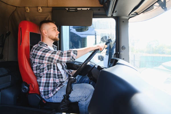 Man sitting in the driver’s seat of a semi-truck cab, holding the steering wheel and looking ahead through the windshield.