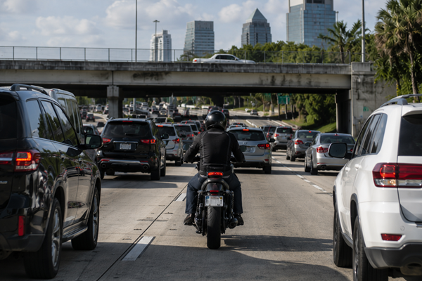 A motorcyclist in a helmet rides through dense freeway traffic surrounded by SUVs and passenger vehicles with the Jacksonville, Florida skyline visible in the background, depicting the dangerous urban riding conditions that contribute to motorcycle accidents in Jacksonville.