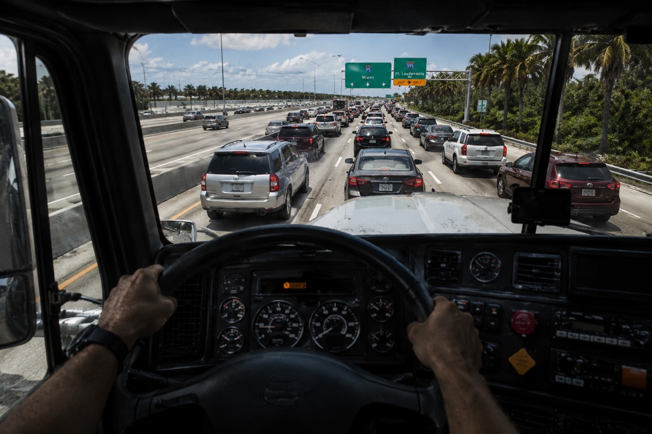 A semi-truck driver's first-person view from the cab, steering through dense highway traffic on Interstate 95 in Florida, as it approaches an exit for Miami and Fort Lauderdale.