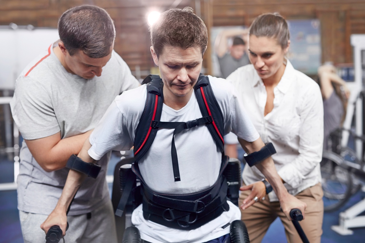 A man using a robotic exoskeleton walking device supported by two therapists during rehabilitation in Florida, illustrating the extensive lifetime treatment costs tied to a spinal cord injury.